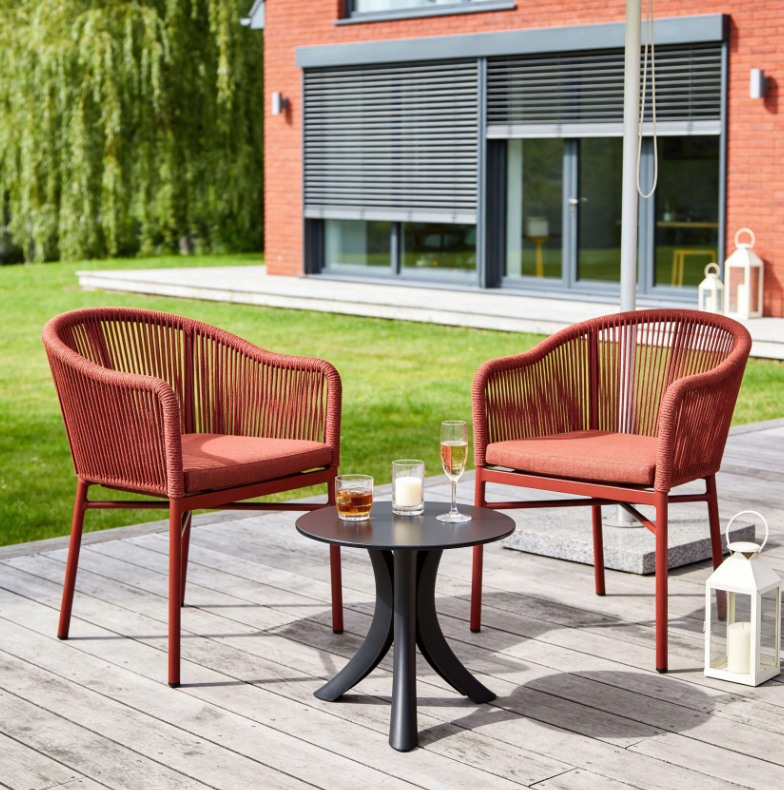 Dwa czerwone, plecione krzesła z poduszkami ustawione naprzeciwko siebie przy małym okrągłym stoliku na drewnianym tarasie.